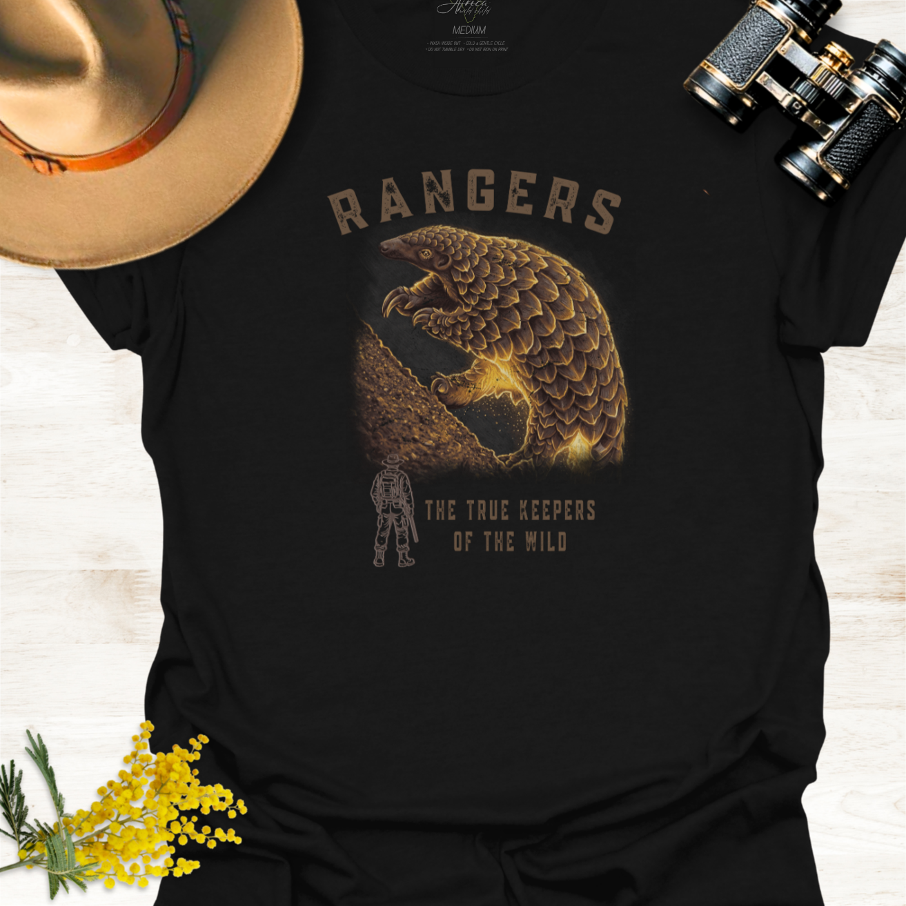 A black graphic T-shirt laid flat on a light wood surface, styled with a tan hat, binoculars, and yellow flowers. The shirt features a large pangolin graphic illustration with the word "RANGERS" above it and the phrase "THE TRUE KEEPERS OF THE WILD" below, plus a small ranger silhouette.