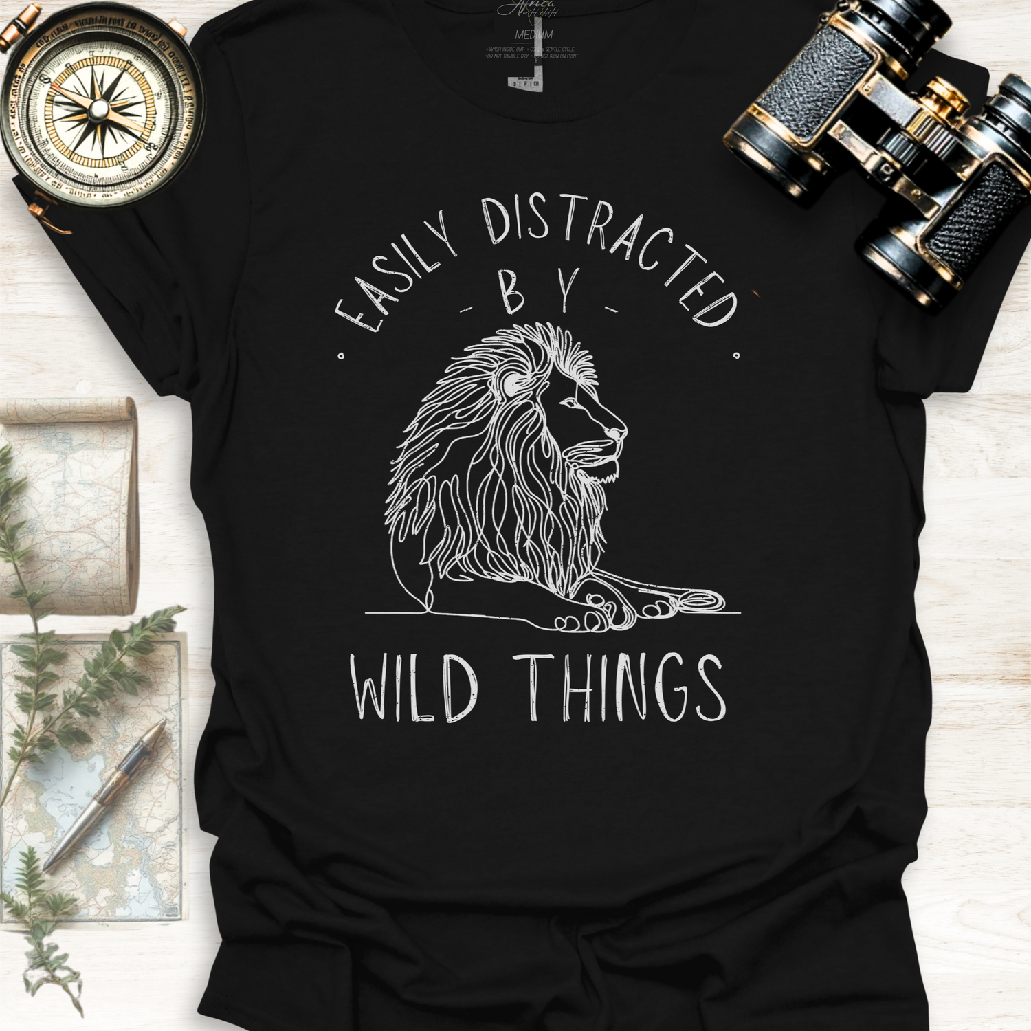 Black t-shirt with a lion graphic and text 'Easily Distracted by Wild Things', with binoculars, a compass, a map and ferns on a light wooden surface.