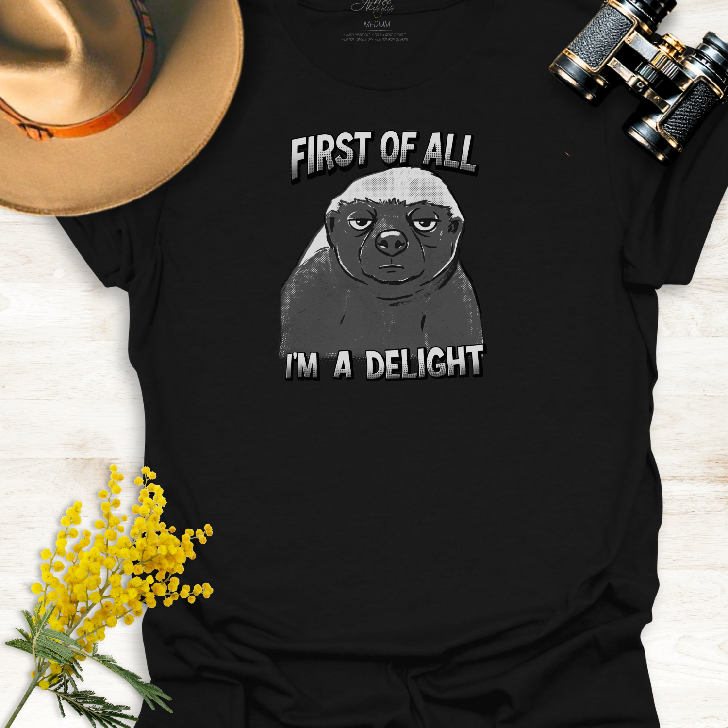 Black t-shirt with a monotone graphic of an unimpressed cartoon illustration of an Africa Honey Badger with caption "First of all I'm a delight" on a wooden surface.