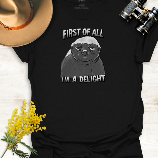 Black t-shirt with a monotone graphic of an unimpressed cartoon illustration of an Africa Honey Badger with caption "First of all I'm a delight" on a wooden surface.