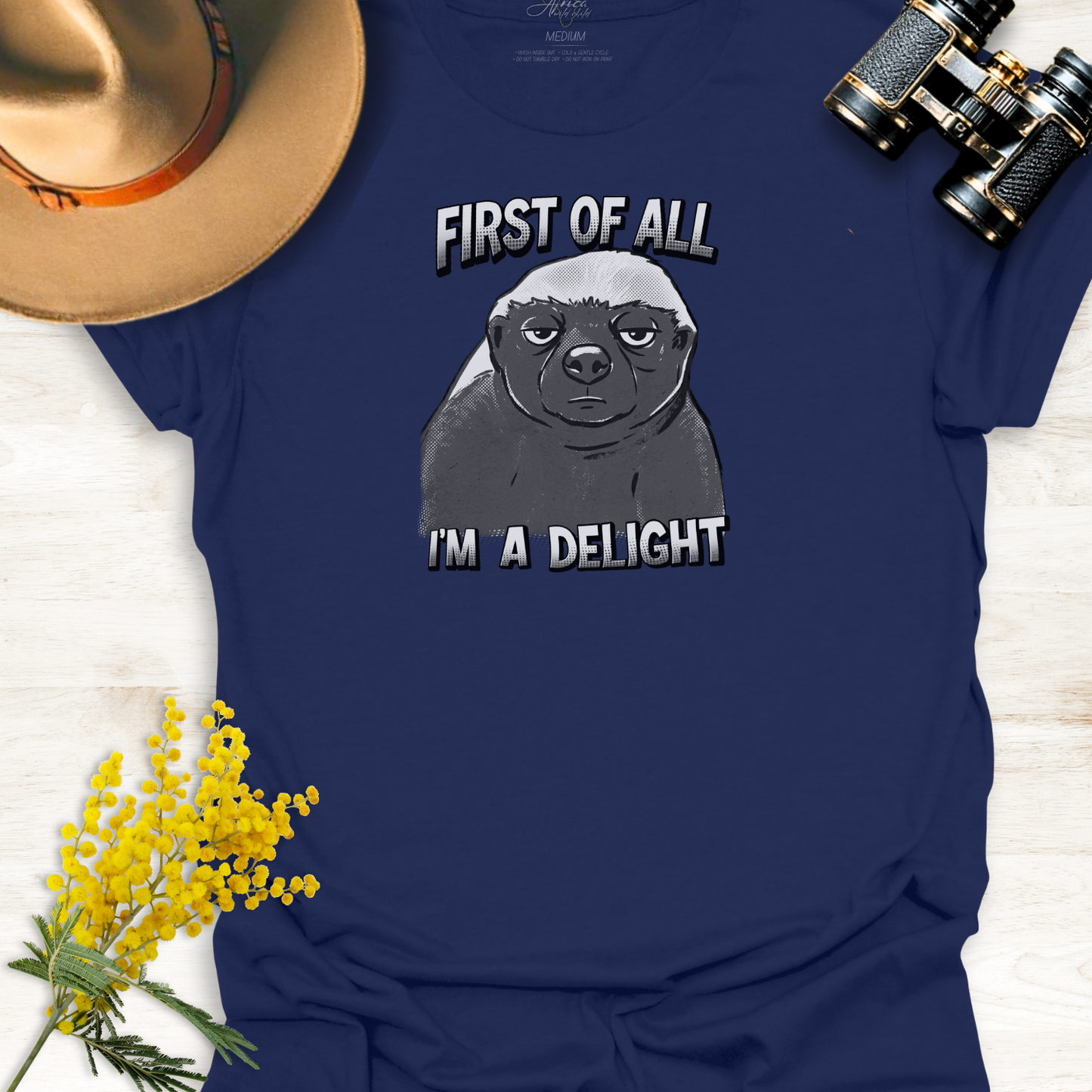 Navy Blue t-shirt with a monotone graphic of an unimpressed cartoon illustration of an Africa Honey Badger with caption "First of all I'm a delight" on a wooden surface.