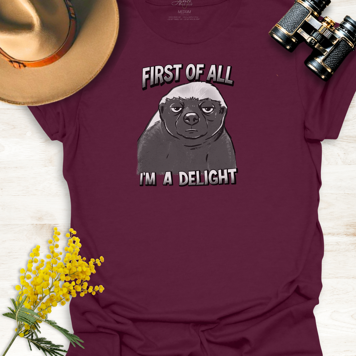 Maroon t-shirt with a monotone graphic of an unimpressed cartoon illustration of an Africa Honey Badger with caption "First of all I'm a delight" on a wooden surface.