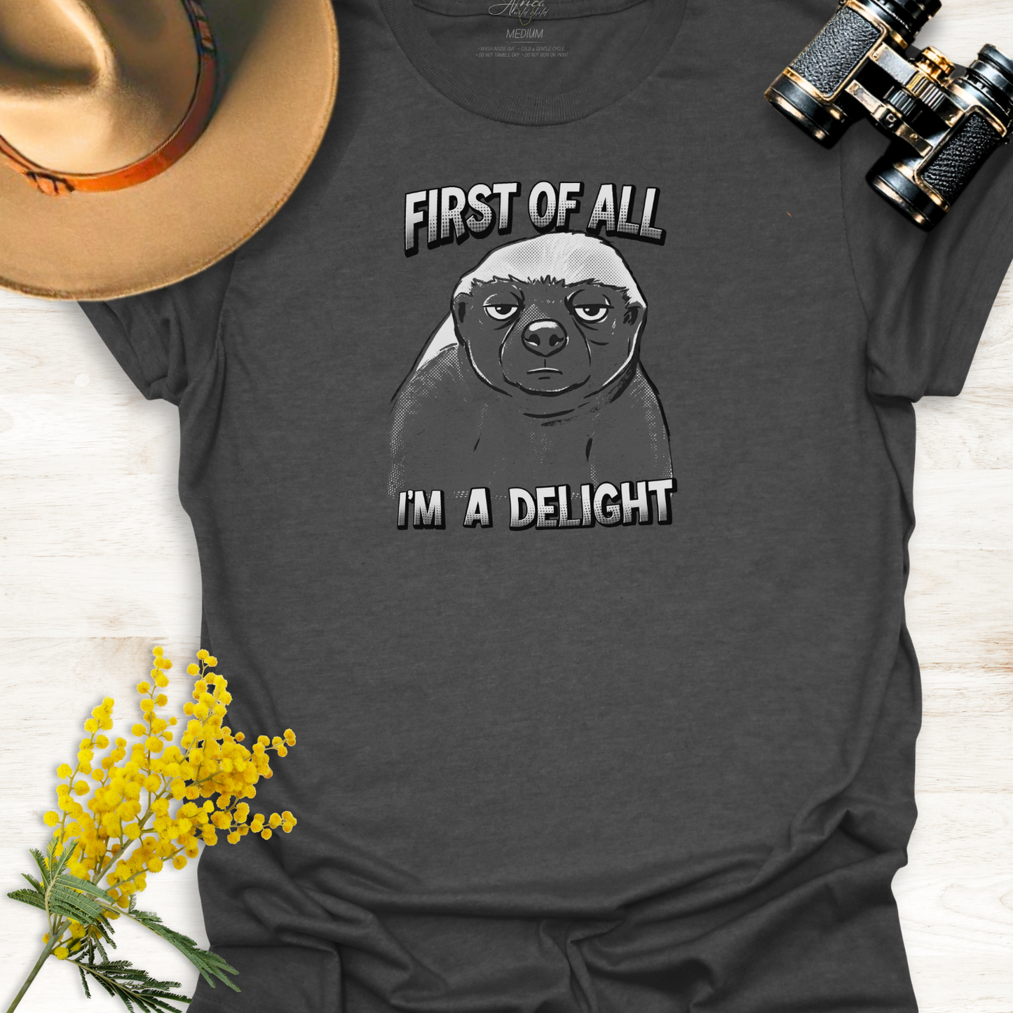 Grey t-shirt with a monotone graphic of an unimpressed cartoon illustration of an Africa Honey Badger with caption "First of all I'm a delight" on a wooden surface.