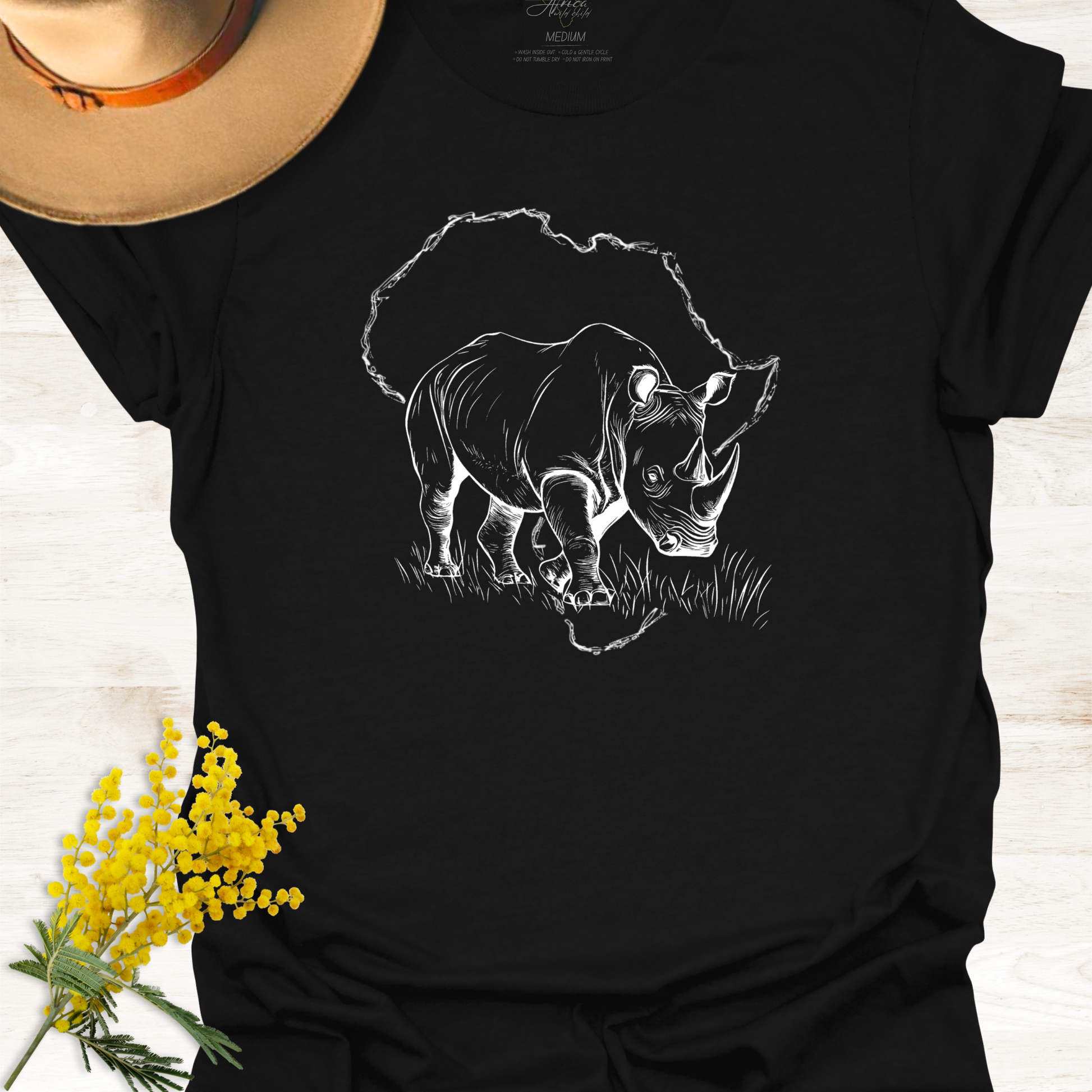 Black t-shirt with a white rhinoceros design on a wooden surface with yellow flowers and a hat.