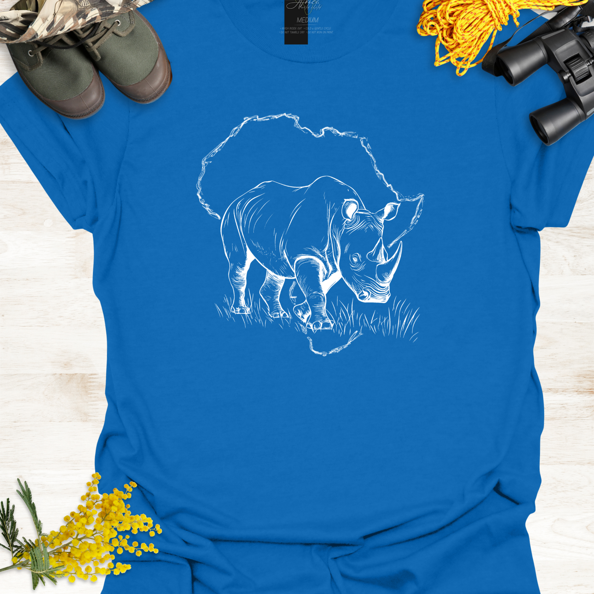Blue t-shirt with a white rhinoceros design on a wooden surface with camouflage boots, yellow flowers, and binoculars.