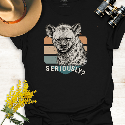 Black t-shirt with an unimpressed hyena graphic illustration and 'SERIOUSLY?' text, surrounded by a hat, binoculars, and yellow flowers on a light wooden surface.