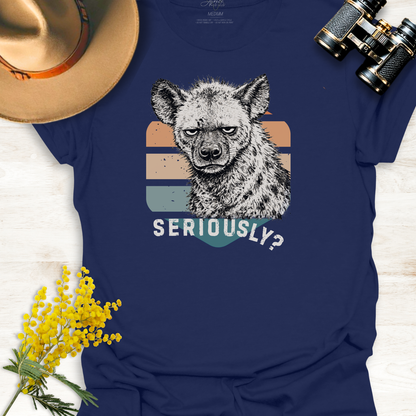 Navy blue t-shirt with an unimpressed hyena graphic illustration and 'SERIOUSLY?' text, surrounded by a hat, binoculars, and yellow flowers on a light wooden surface.