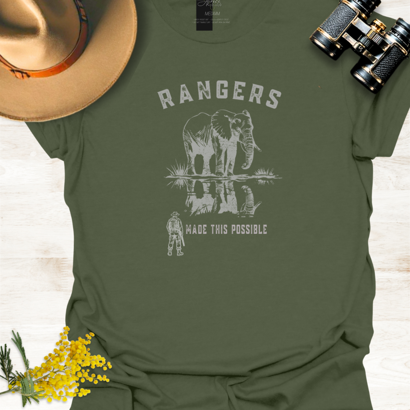 An military green graphic t-shirt featuring a white print of an elephant and a ranger with the text 'RANGERS MADE THIS POSSIBLE'. The shirt is styled in a flat-lay arrangement with a tan hat, black binoculars, and yellow flowers on a light wood background.