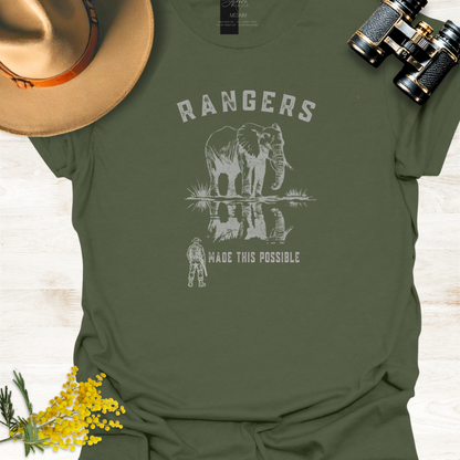 An military green graphic t-shirt featuring a white print of an elephant and a ranger with the text 'RANGERS MADE THIS POSSIBLE'. The shirt is styled in a flat-lay arrangement with a tan hat, black binoculars, and yellow flowers on a light wood background.