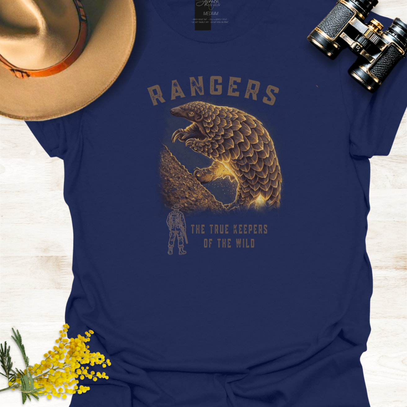 A navy blue graphic T-shirt laid flat on a light wood surface, styled with a tan hat, binoculars, and yellow flowers. The shirt features a large pangolin graphic illustration with the word 'RANGERS' above and the phrase 'THE TRUE KEEPERS OF THE WILD' below.