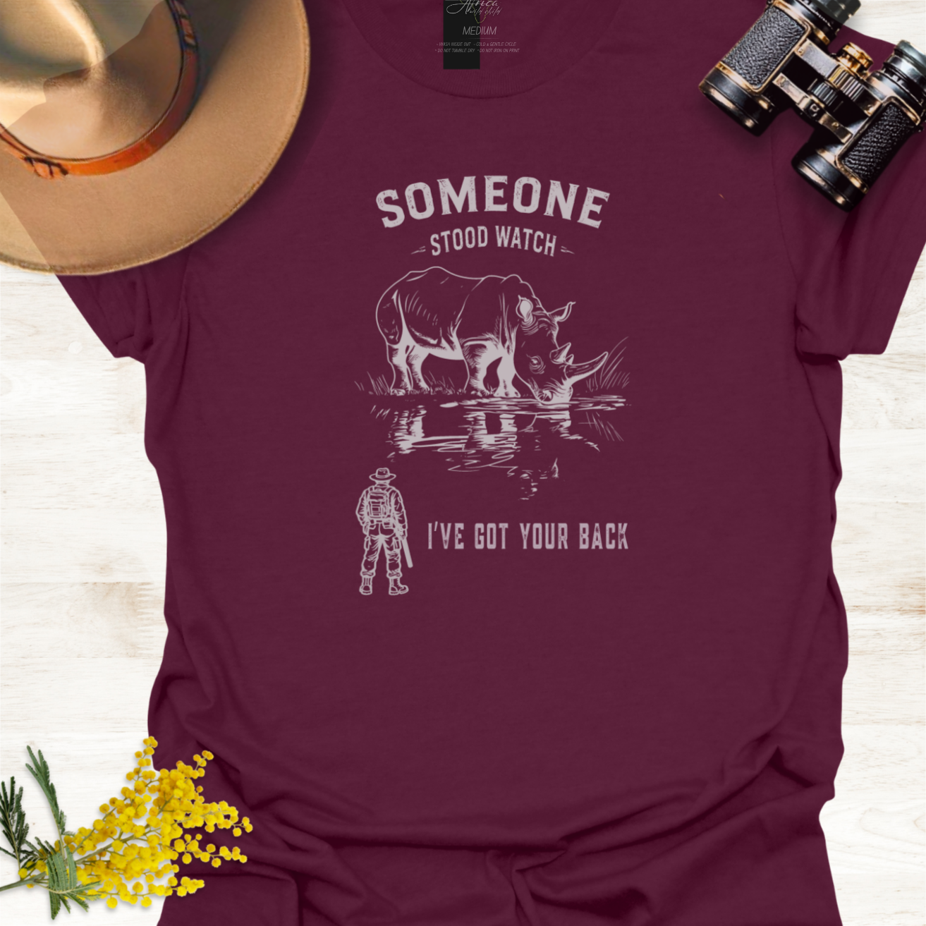 A maroon short-sleeve graphic T-shirt laid flat on a light wood surface, styled with a tan hat, black binoculars, and small yellow flowers. The shirt has a white graphic of a rhinoceros and a ranger with the text 'SOMEONE STOOD WATCH' and 'I'VE GOT YOUR BACK.