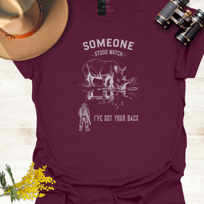 A maroon short-sleeve graphic T-shirt laid flat on a light wood surface, styled with a tan hat, black binoculars, and small yellow flowers. The shirt has a white graphic of a rhinoceros and a ranger with the text 'SOMEONE STOOD WATCH' and 'I'VE GOT YOUR BACK.