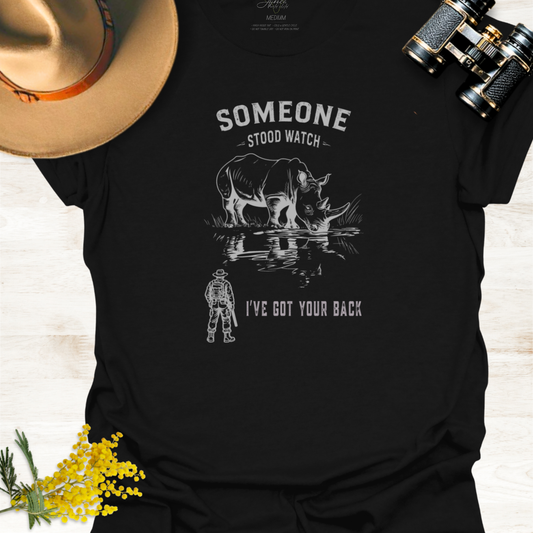 A black graphic t-shirt laid flat on a light wood background, styled with a hat, binoculars, and yellow flowers. The shirt features a white graphic of a rhinoceros and a ranger with the text 'SOMEONE STOOD WATCH I'VE GOT YOUR BACK'.