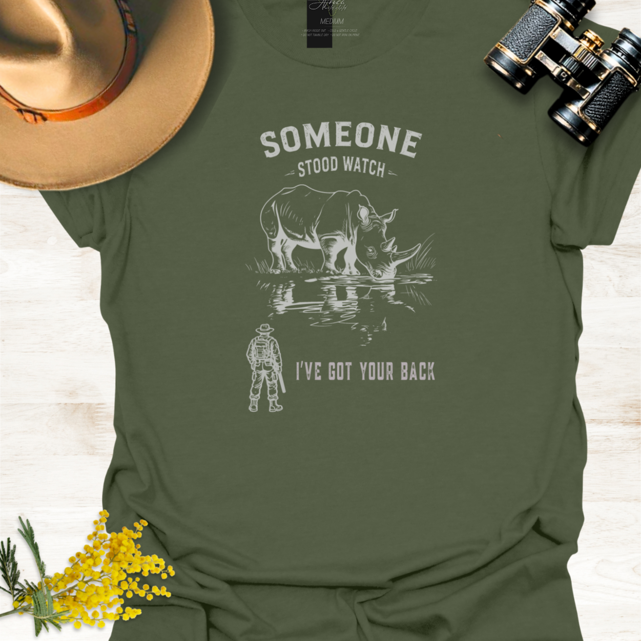 An military green short-sleeve graphic T-shirt laid flat on a light wood surface, styled with a tan hat, black binoculars, and small yellow flowers. The shirt has a white graphic of a rhinoceros and a ranger with the text 'SOMEONE STOOD WATCH' and 'I'VE GOT YOUR BACK.