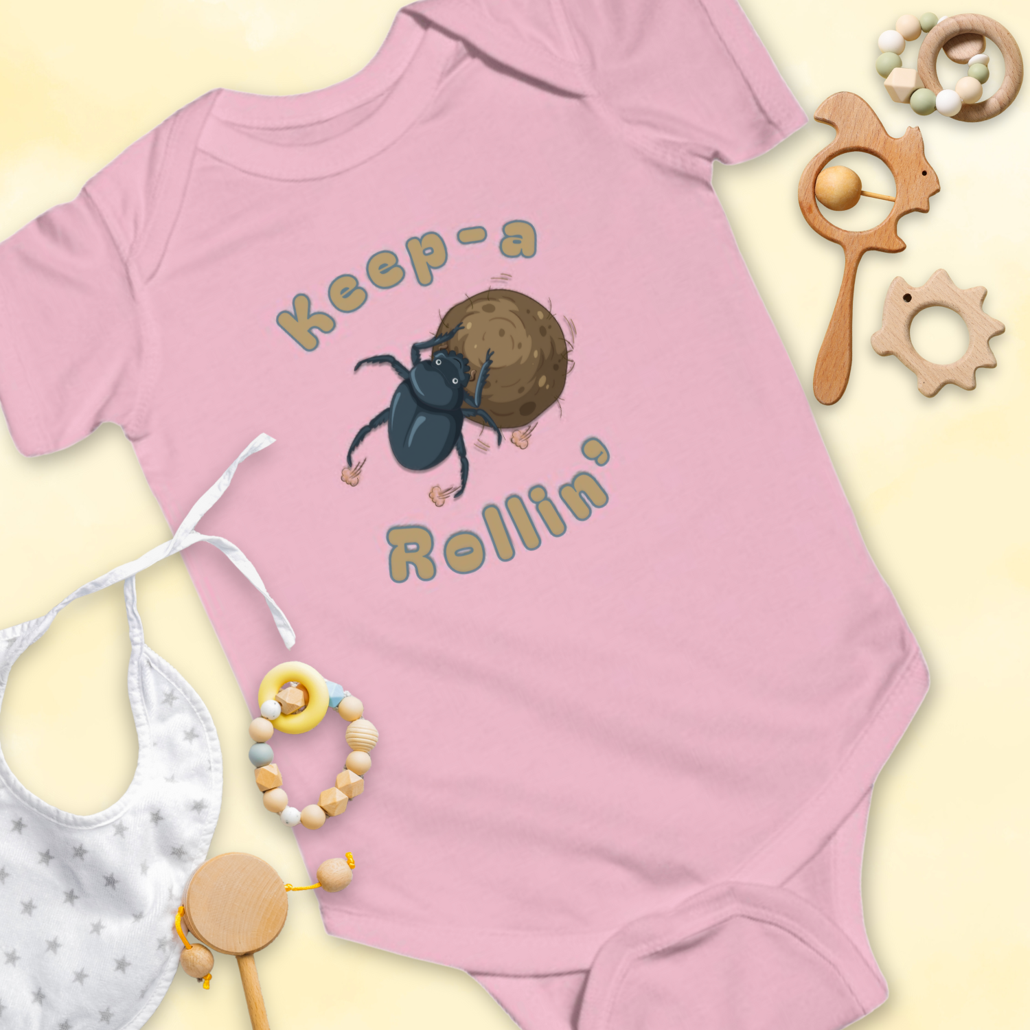 Pink baby onesie with dung beetle graphic and 'Keep-a Rollin'' text, surrounded by baby items on a light 
