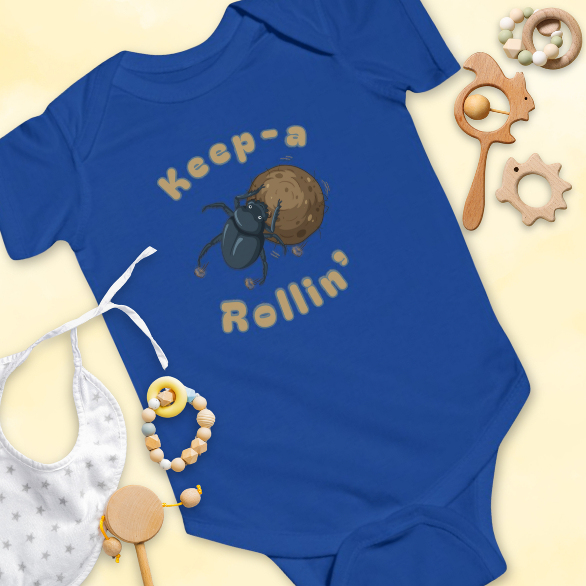 Blue baby onesie with dung beetle graphic and 'Keep-a Rollin'' text, surrounded by baby items on a light 