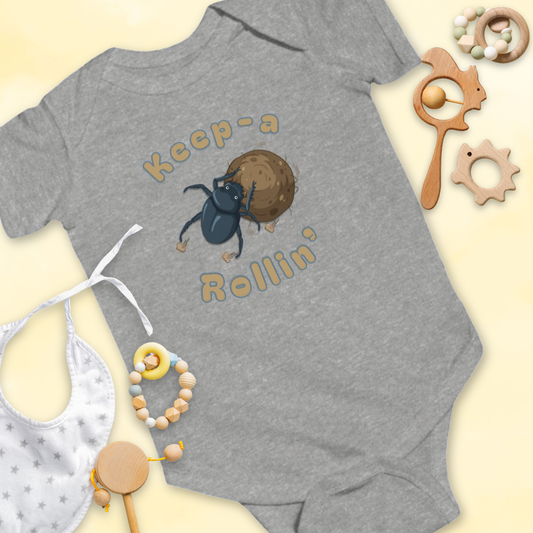 Gray baby onesie with dung beetle graphic and 'Keep-a Rollin'' text, surrounded by baby items on a light yellow background.