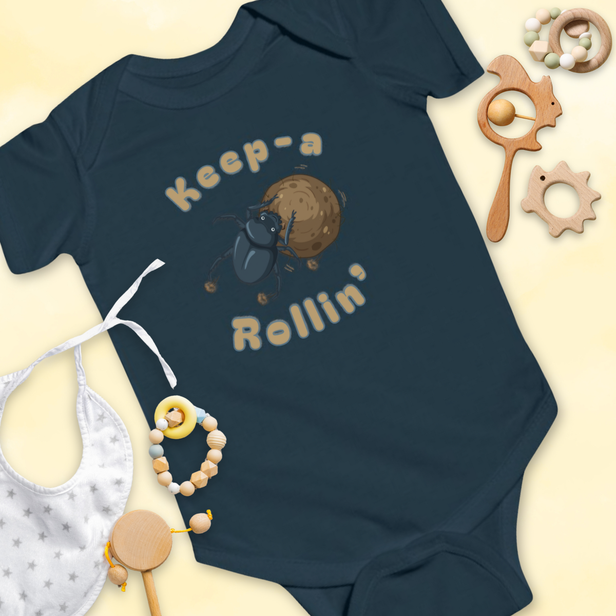 Navy blue baby onesie with dung beetle graphic and 'Keep-a Rollin'' text, surrounded by baby items on a light 