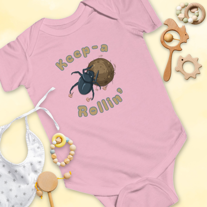 Pink baby onesie with dung beetle graphic and 'Keep-a Rollin'' text, surrounded by baby items on a light 