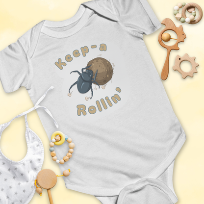 Gray baby onesie with dung beetle graphic and 'Keep-a Rollin'' text, surrounded by baby items on a light 