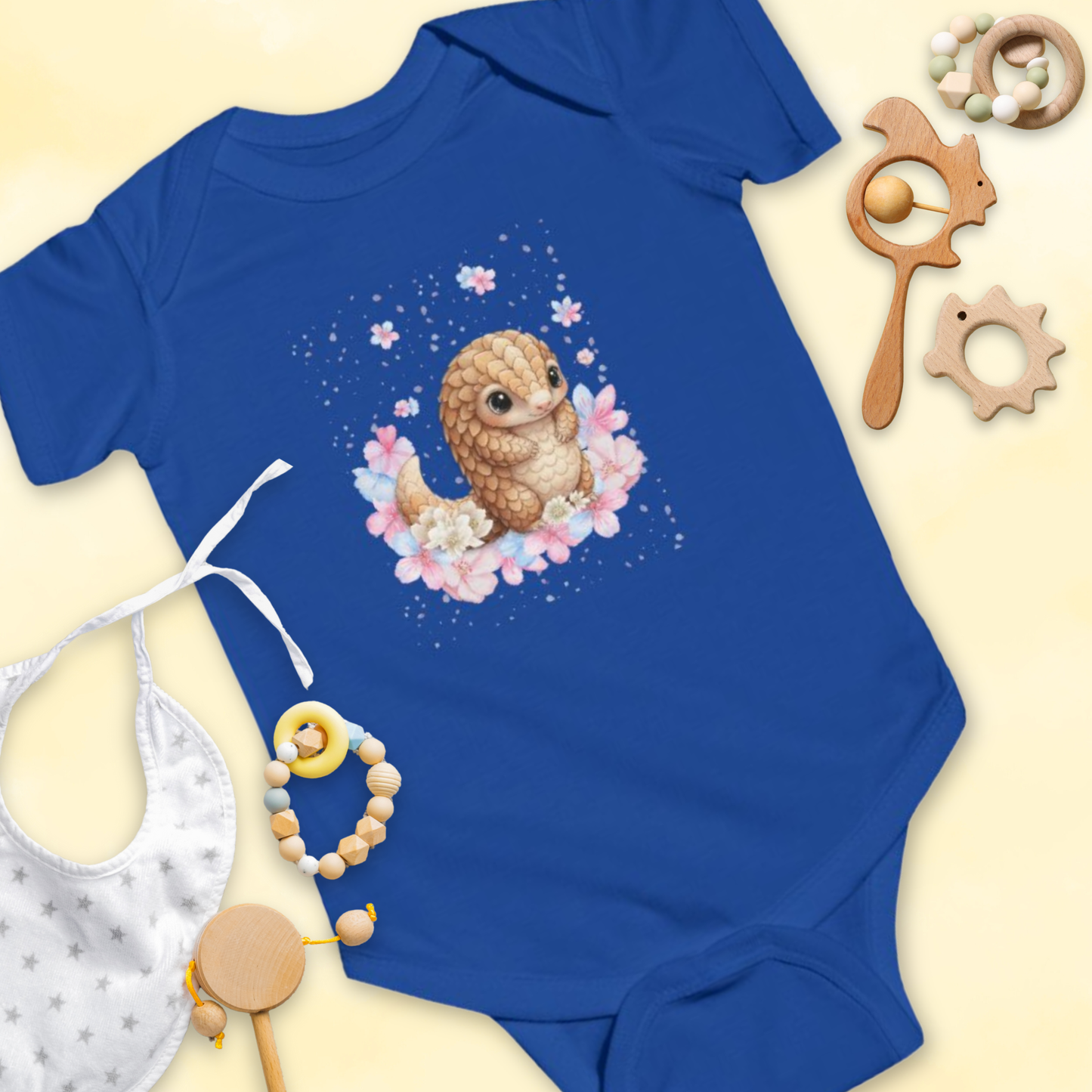 Blue baby onesie with a cartoon pangolin & flower design, surrounded by baby toys on a light yellow background.