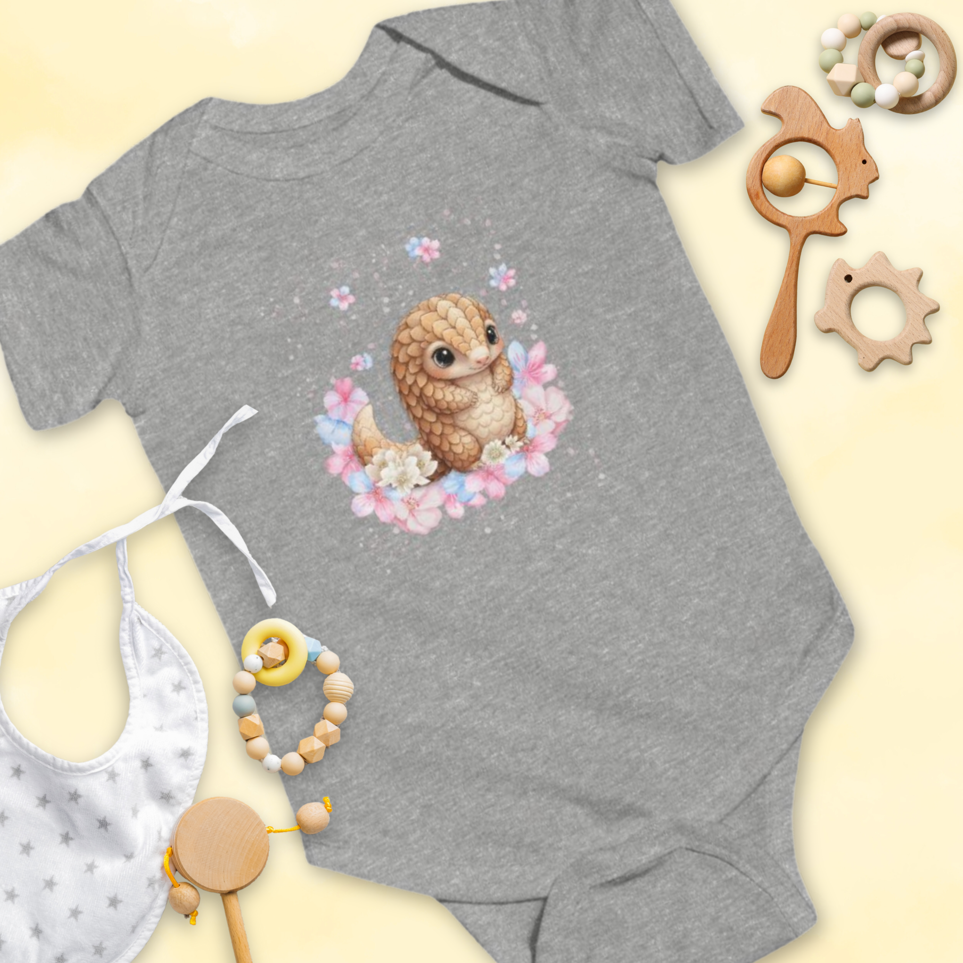Gray baby onesie with a cartoon pangolin & flower design, surrounded by baby toys on a light yellow background.