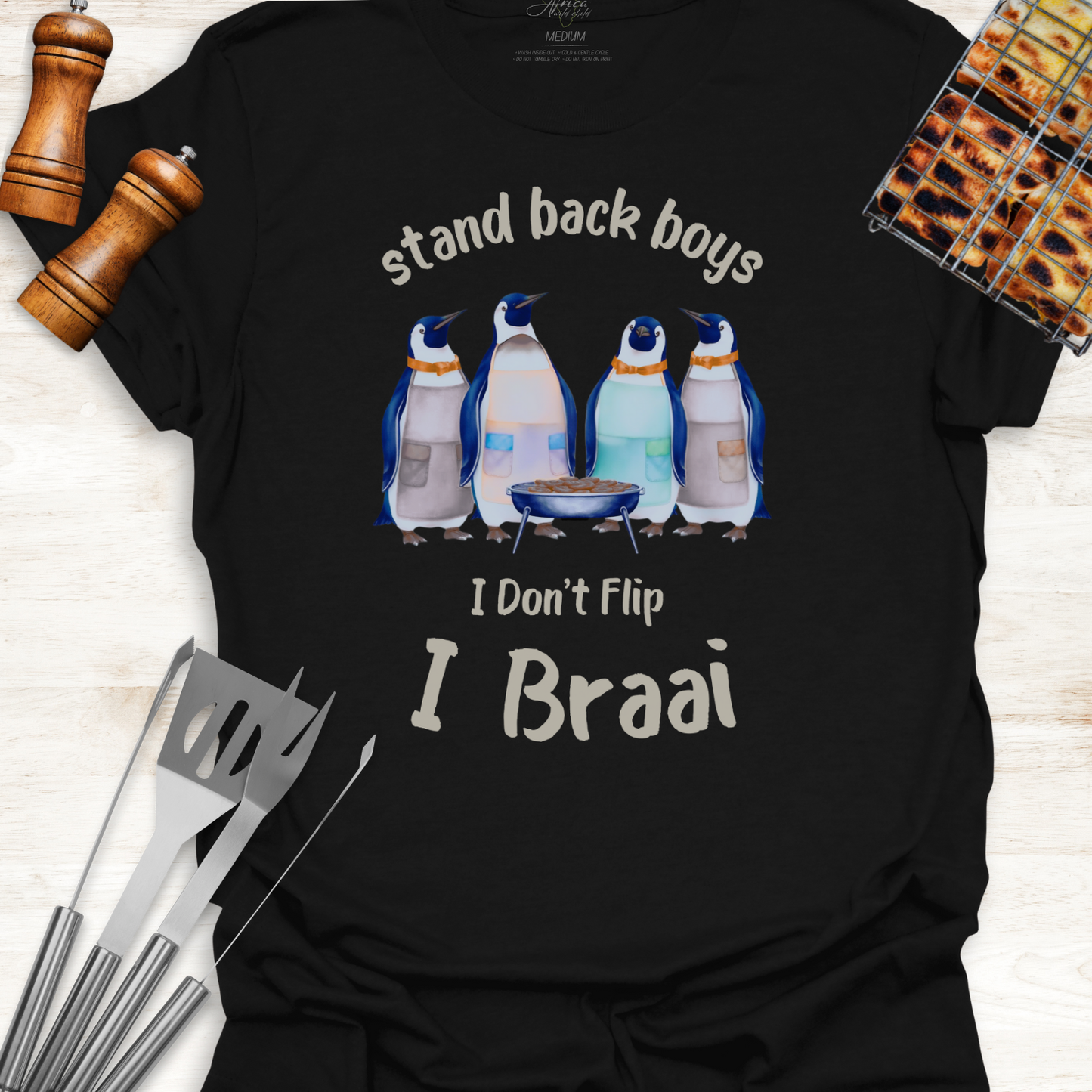 Black t-shirt with penguin graphic and text 'stand back boys I Don't Flip I Braai' on a wooden surface with grill tools.