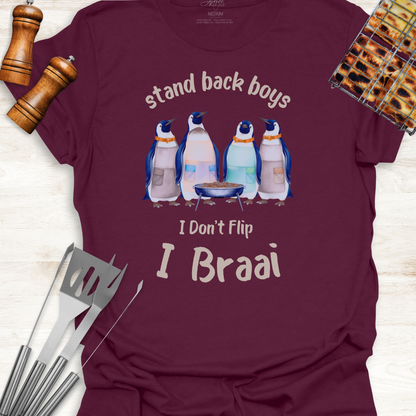 Maroon t-shirt with penguin graphic and text 'stand back boys, I don't flip, I Braai' on a wooden surface with grill tools.