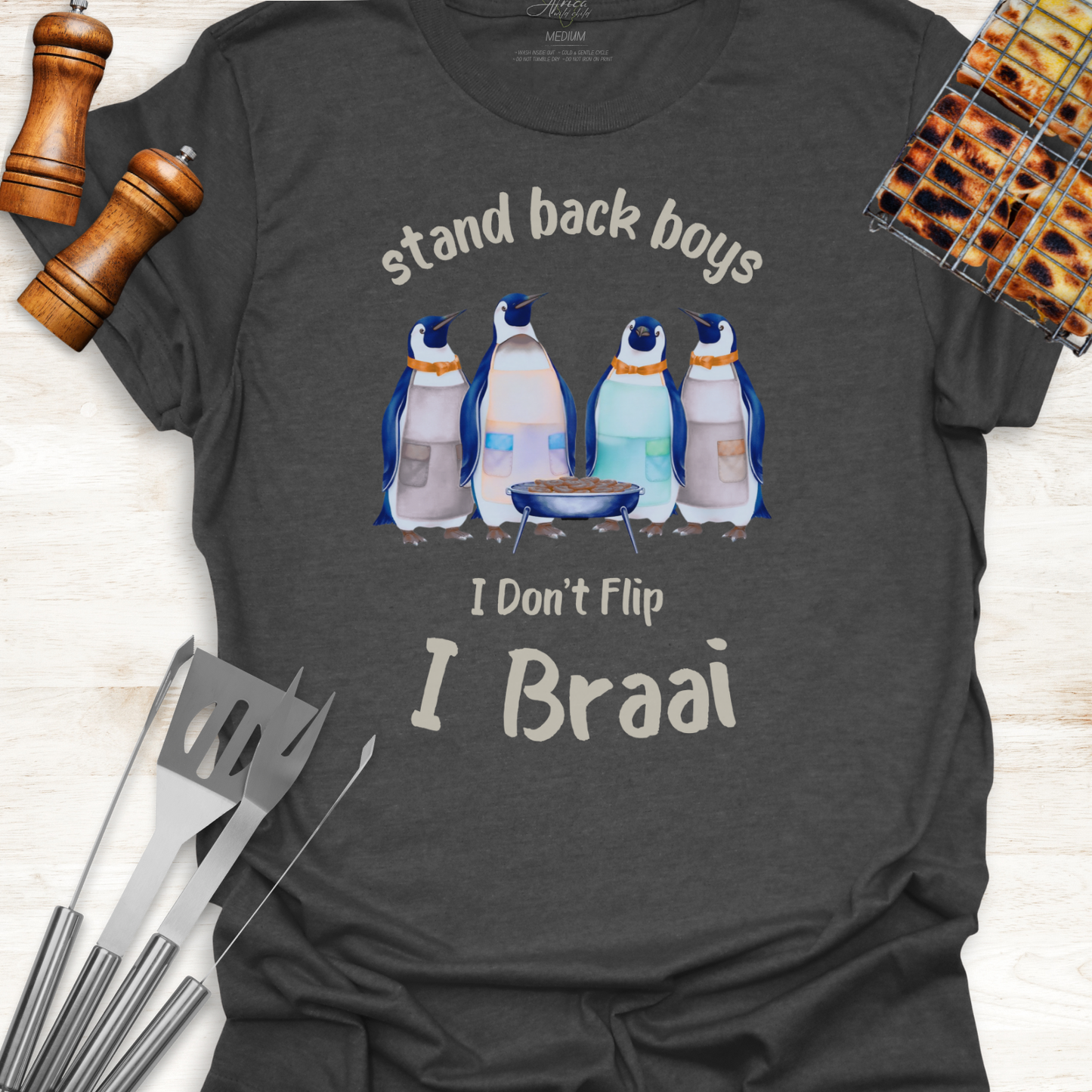 Dark gray t-shirt with penguin graphic and text 'stand back boys, I Don't Flip I Braai' on a wooden surface with grill tools.