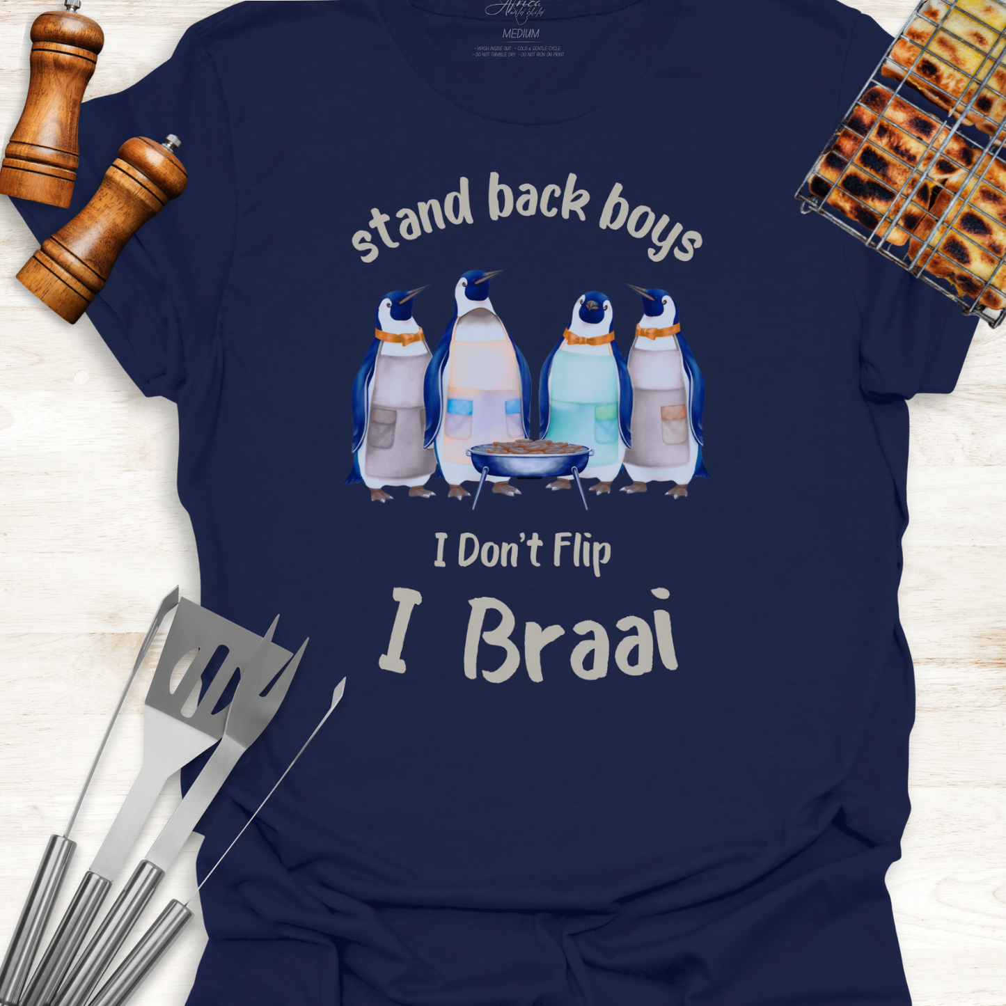 Navy blue t-shirt with penguin graphic and text 'stand back boys, I Don't Flip, I Braai' on a wooden surface with grill tools.