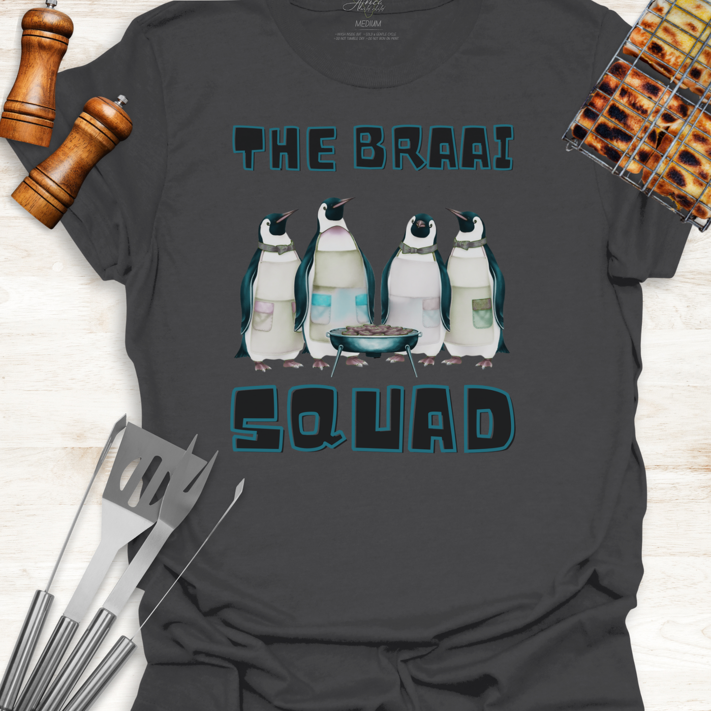 Gray t-shirt with penguin design and text 'The Braai Squad' on a wooden surface with grill tools.