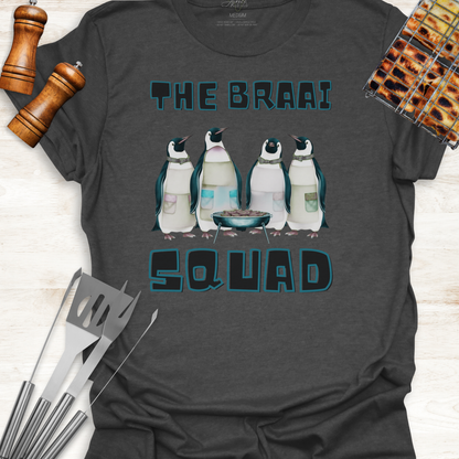Gray t-shirt with penguin graphic and 'The Braai Squad' text on a wooden surface with grill tools.