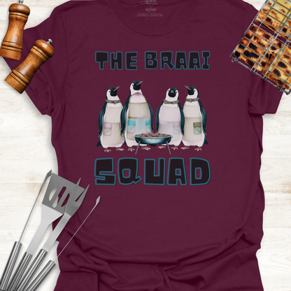 Maroon t-shirt with penguin graphic and 'The Braai Squad' text, surrounded by grill tools and a pepper grinder.