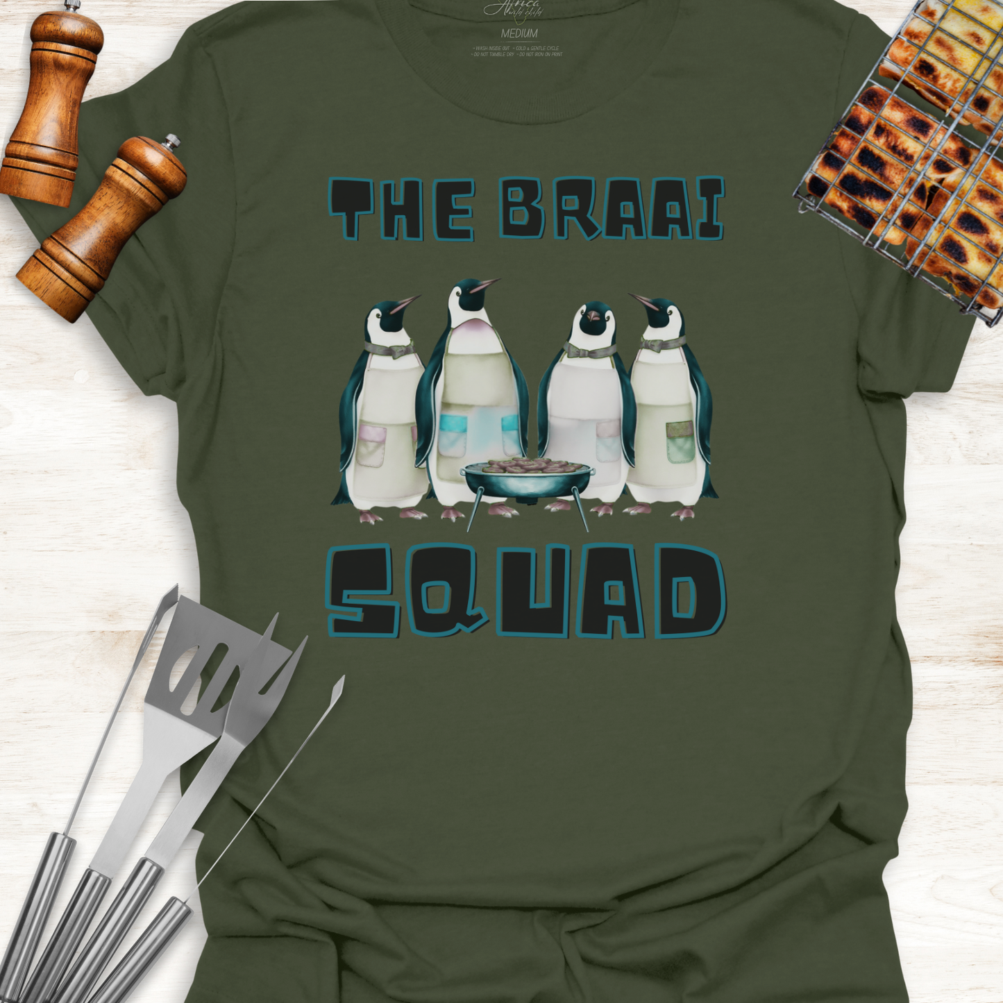 Green t-shirt with penguin design and text 'The Braai Squad' on a wooden surface with grill tools.