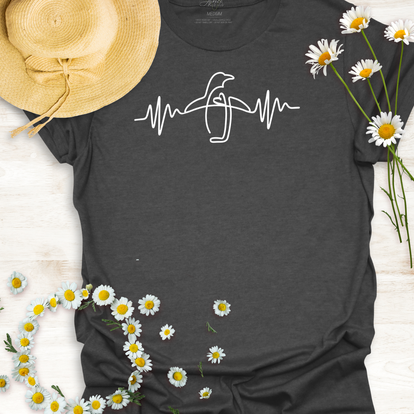 Dark gray wildlife t-shirt with a penguin heartbeat design, surrounded by daisies and a straw hat on a light wooden surface.
