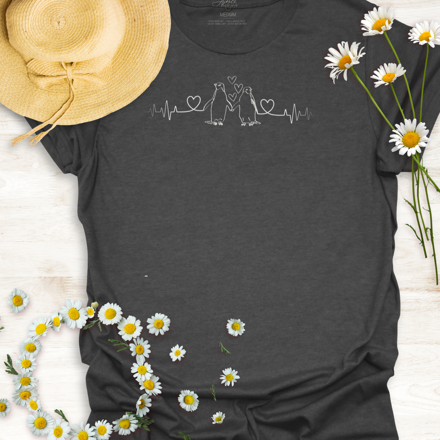 Dark gray  t-shirt with hearts on a heartbeat line with penguins design, surrounded by daisies and a straw hat on a light wooden surface.