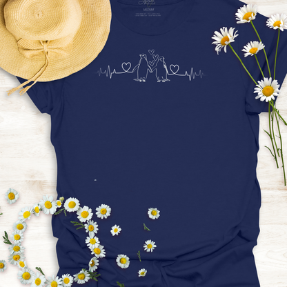 Navy blue  t-shirt with hearts on a heartbeat line with penguins design, surrounded by daisies and a straw hat on a light wooden surface.