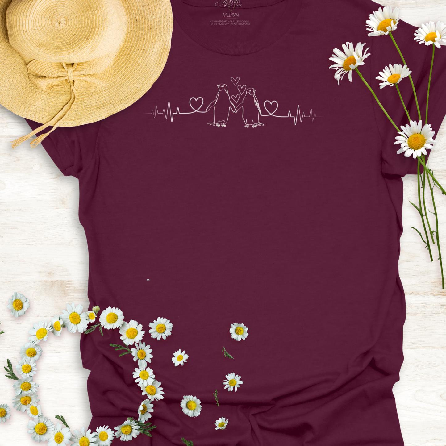 Maroon t-shirt with hearts on a heartbeat line with penguins design, surrounded by daisies and a straw hat on a light wooden surface.