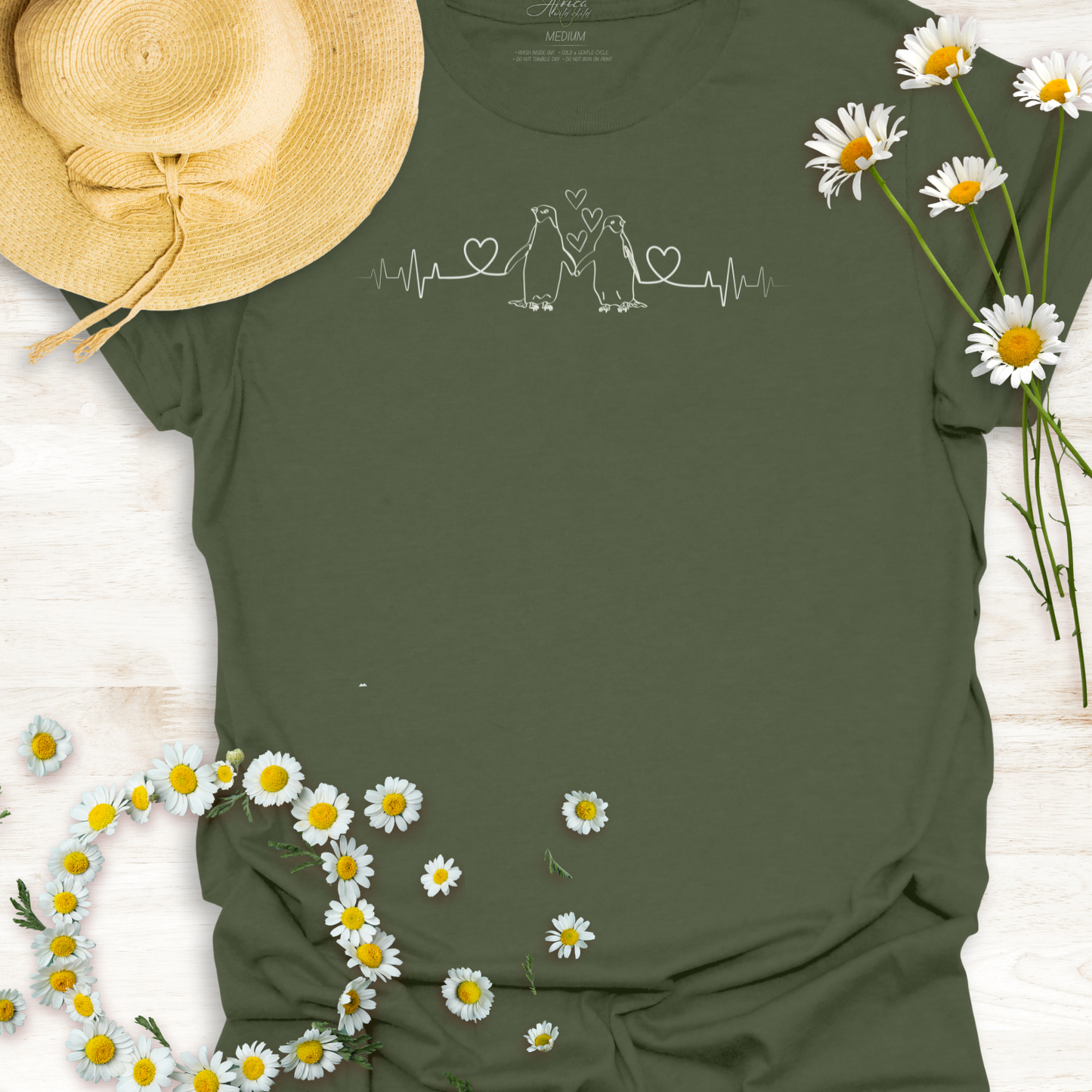 Green t-shirt with hearts on a heartbeat line with penguins print design, surrounded by daisies and a straw hat on a light wooden surface.
