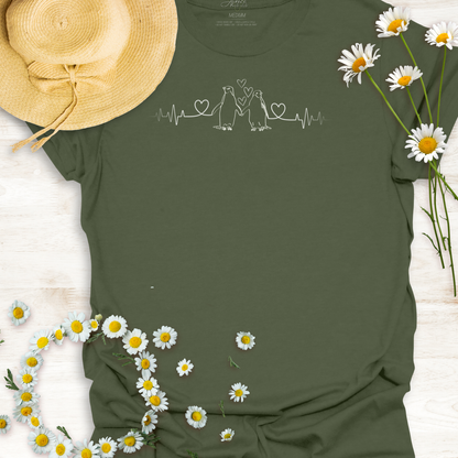 Green t-shirt with hearts on a heartbeat line with penguins print design, surrounded by daisies and a straw hat on a light wooden surface.